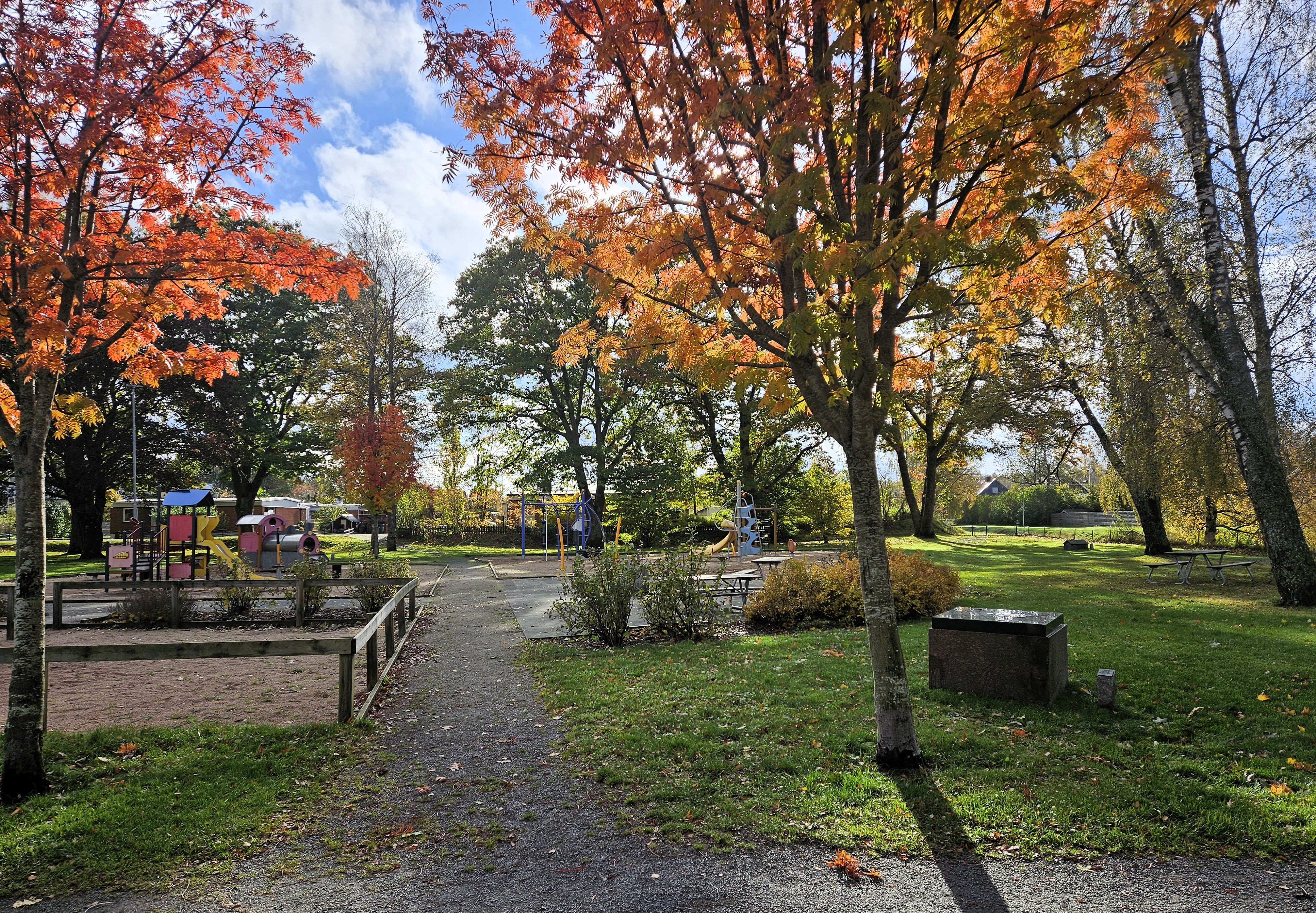 En lekplats ligger inbäddad i grönska under hösten. Färgerna på träden är varma hösttoner, och fler