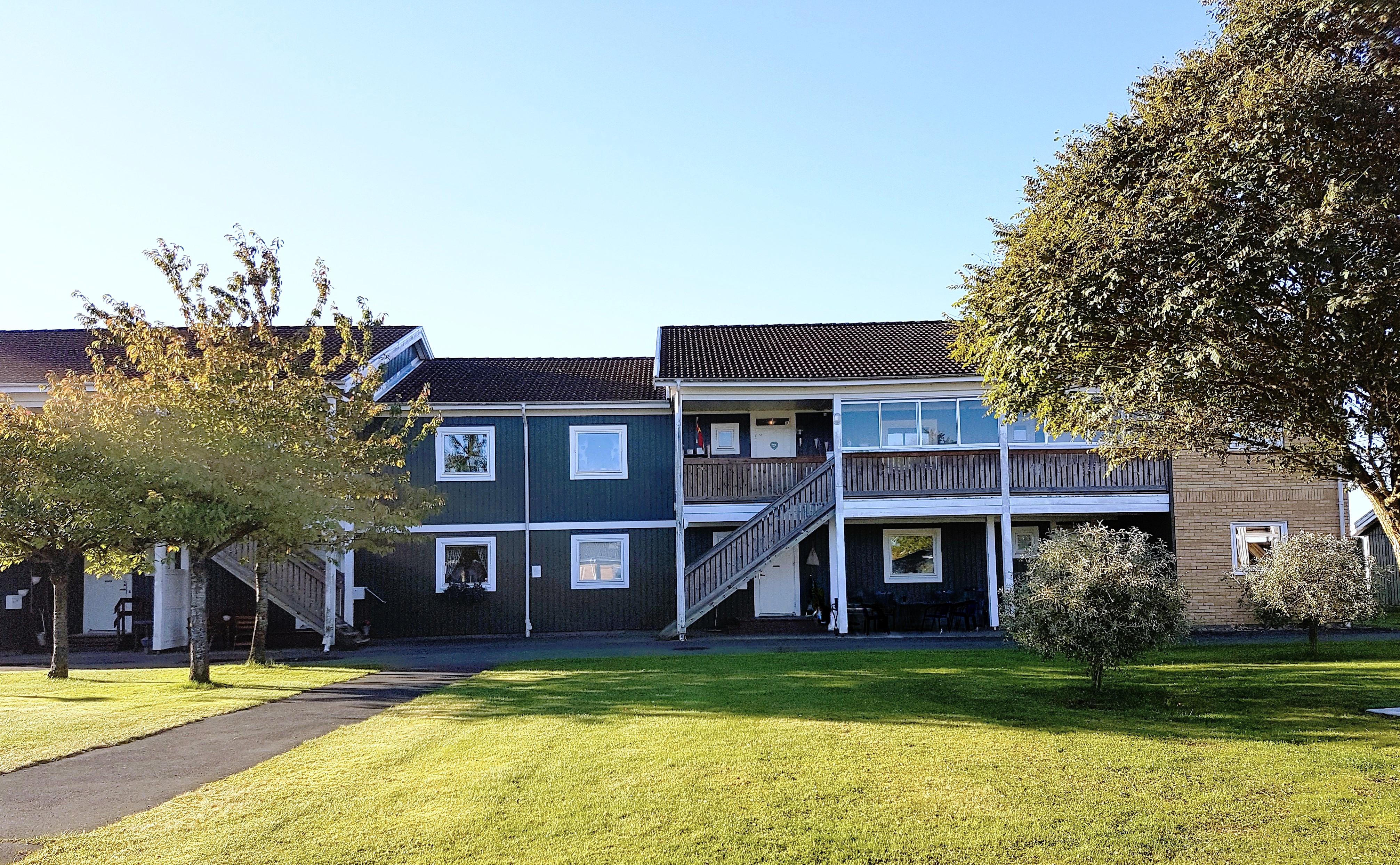 Hus med två våningar i grönt trä. I förgrunden är det träd och en gräsmatta